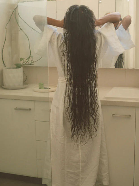 Person with long hair standing in a bathroom, wearing a white robe.