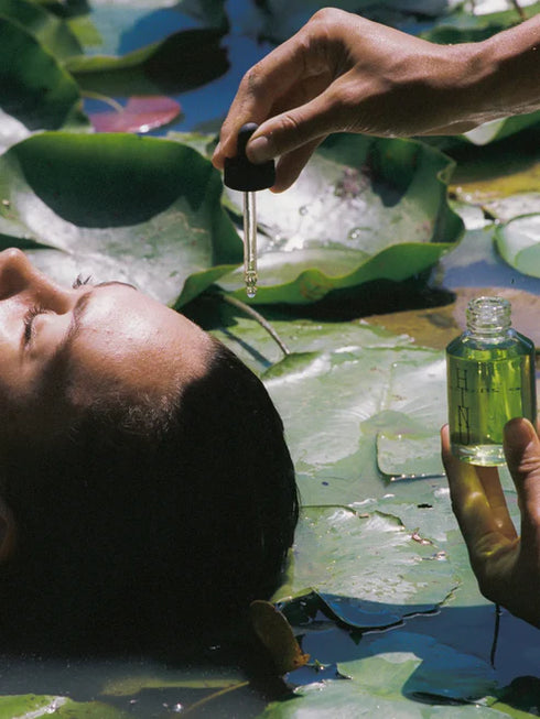 Person lying on water lilies with another hand holding a bottle of green liquid.