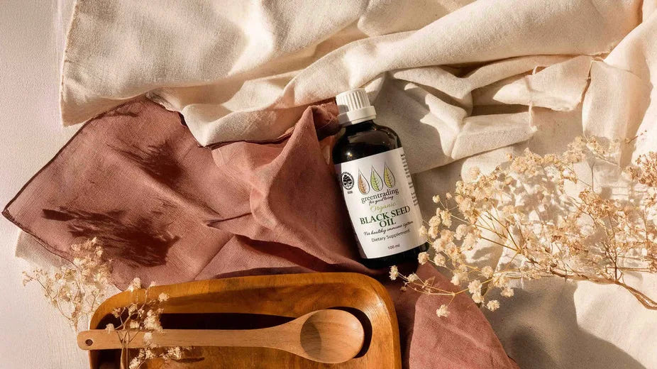 Green Trading Glass Bottle Black Seed Oil on a flat lay image surrounded by earthy tone cloth and flowers
