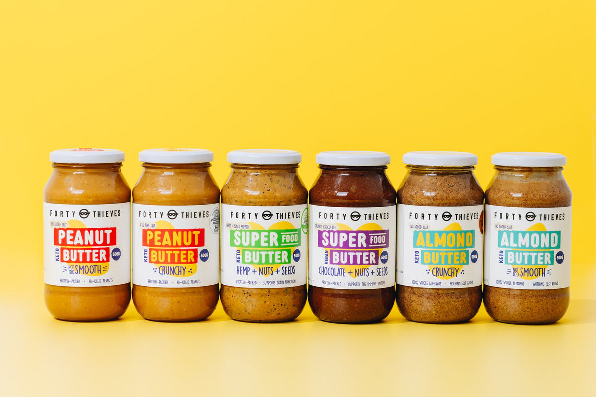 Six 500g jars of Forty Thieves nut butter on a yellow background: peanut smooth, peanut crunchy, hemp super food, chocolate super food, almond crunchy and almond smooth