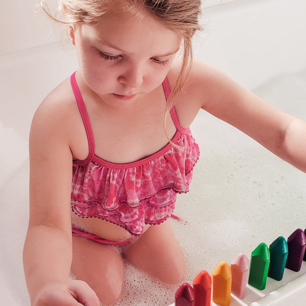 A child in the bath reaching for a tray of coloured bath crayons