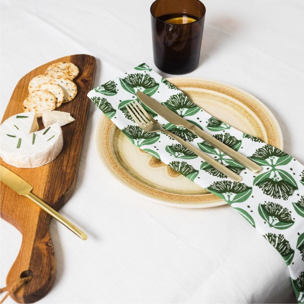 The pohutukawa napkins in shades of green on a set table with a wooden board
