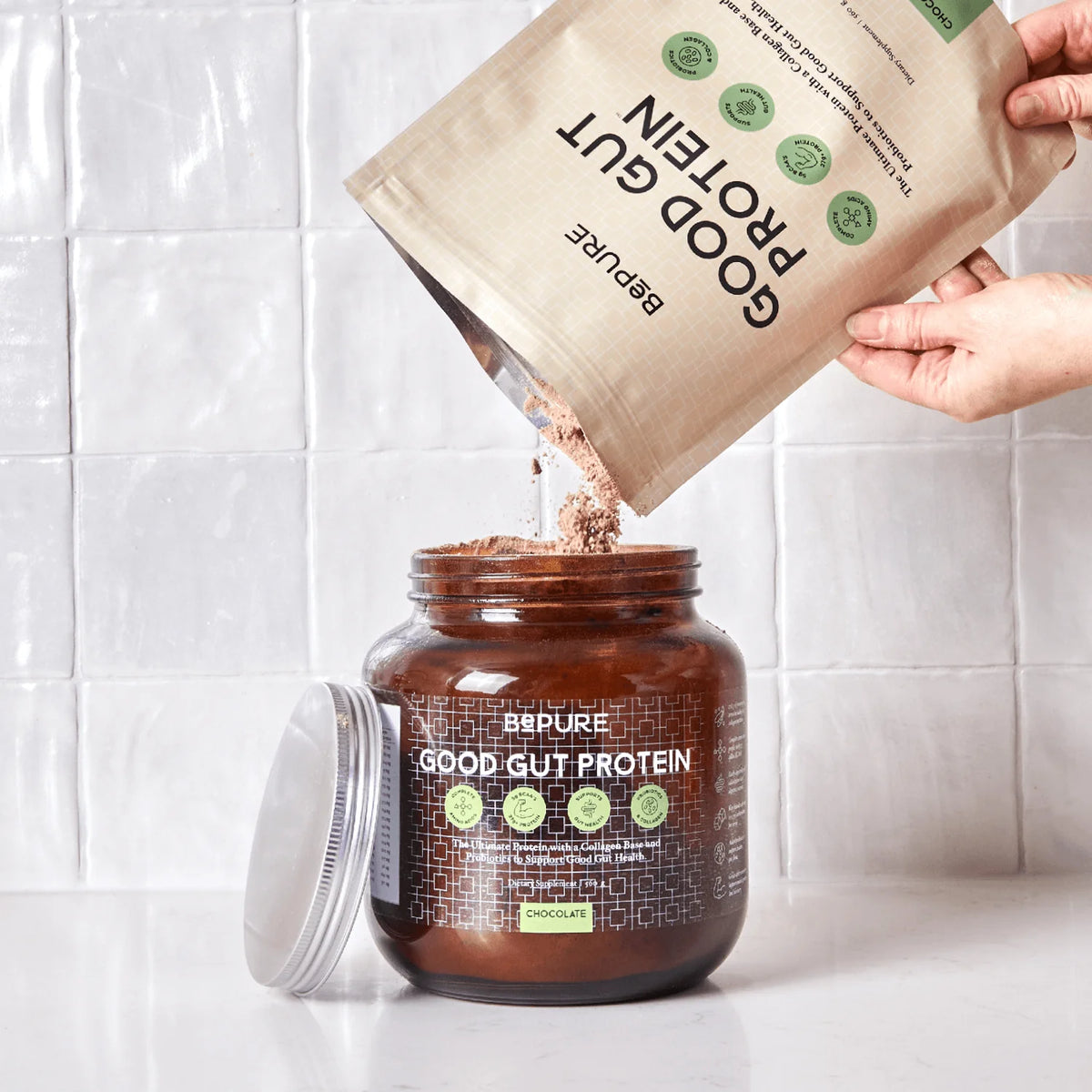 Person pouring protein powder from a bag into a container labeled 'Good Gut Protein' on a white tiled background.