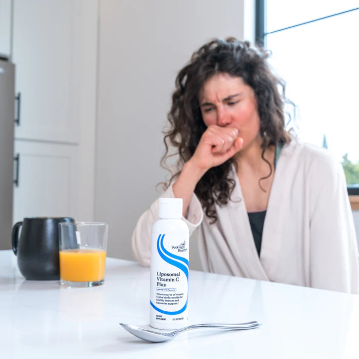 Woman sitting at a table with a bottle of Liposomal Vitamin C Plus, a glass of orange juice, and a spoon.