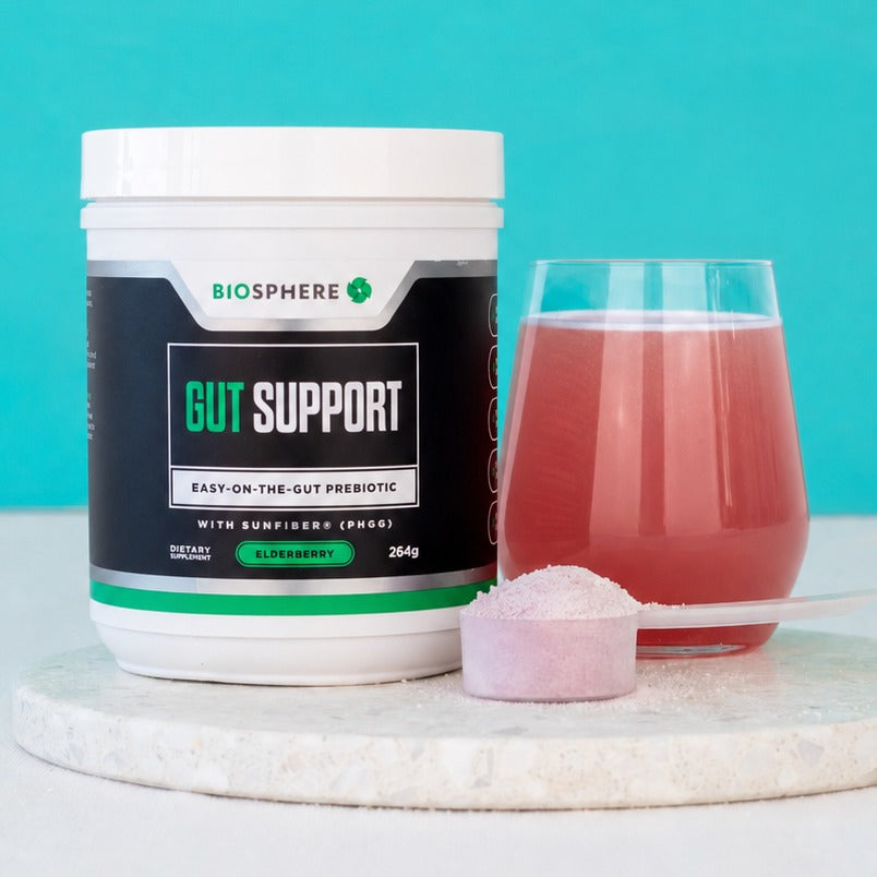 Biosphere Gut Support tub on a marble board beside a glass of mixed elderberry drink and a scoop of pink powder