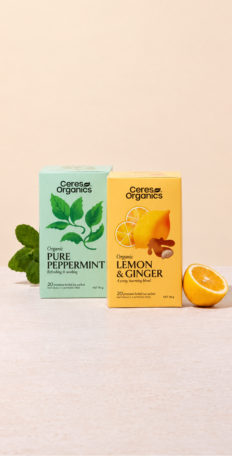 Two Ceres Organics tea boxes on a light beige surface against a warm cream background: a mint green Pure Peppermint box on the left with green leaf graphics, and a yellow Lemon & Ginger box on the right with orange and ginger illustrations. A mint leaf sprig is beside the left box, and a halved orange sits beside the right box.