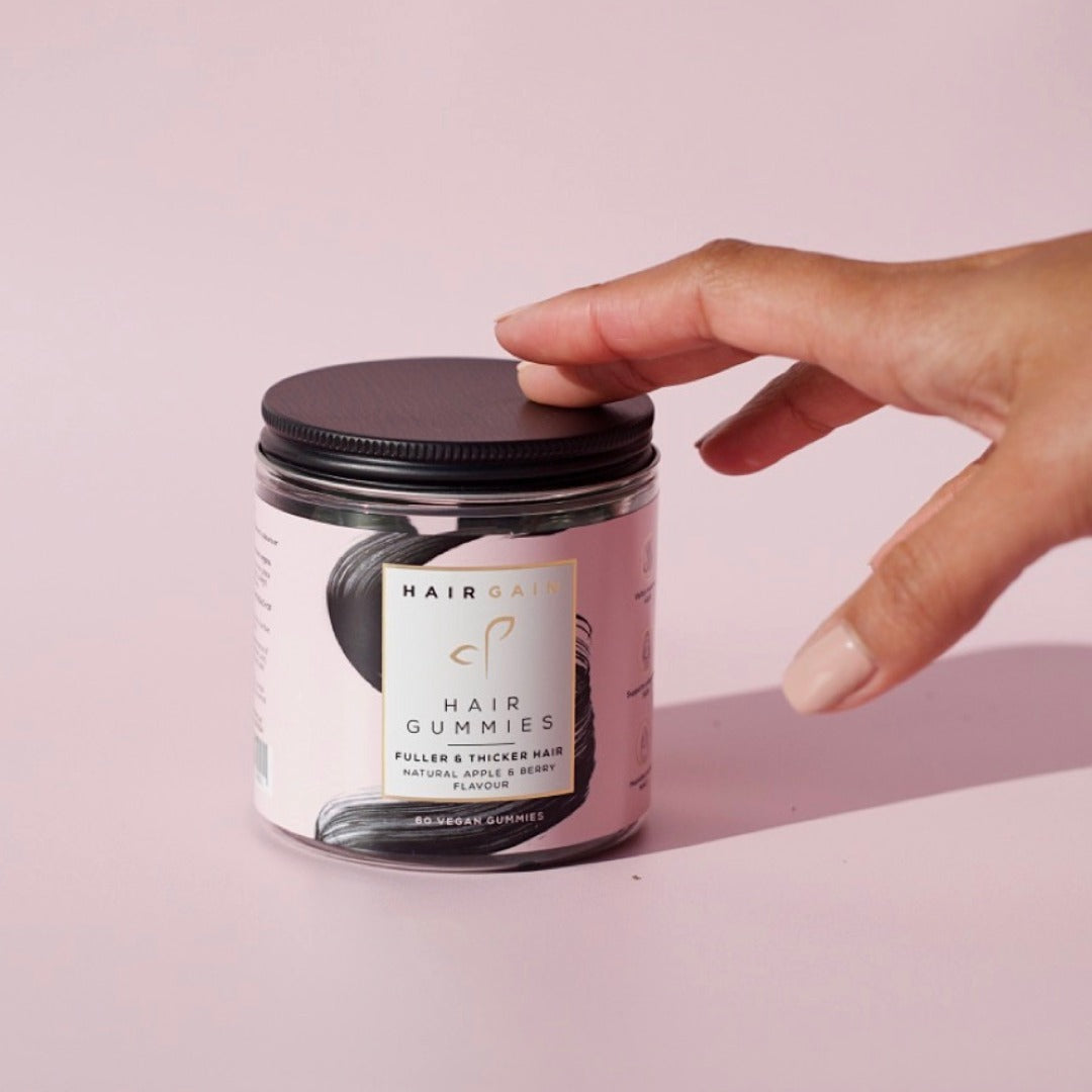 Hand lifting the lid of a Hair Gain Hair Gummies jar on a pink background