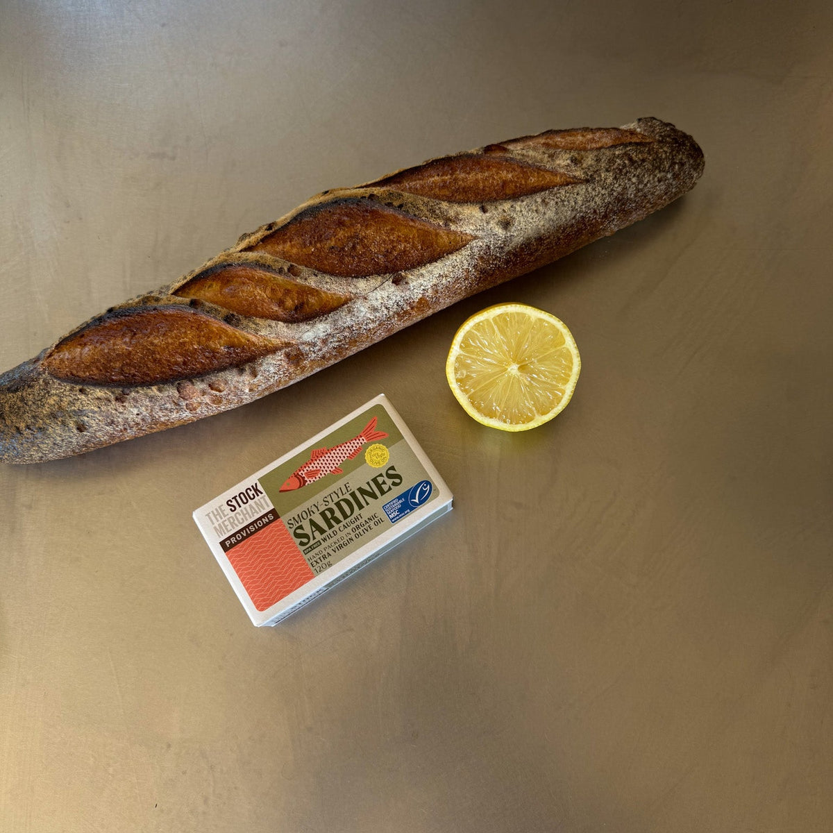 Open tin of smoky style sardines beside a sliced baguette and a lemon half