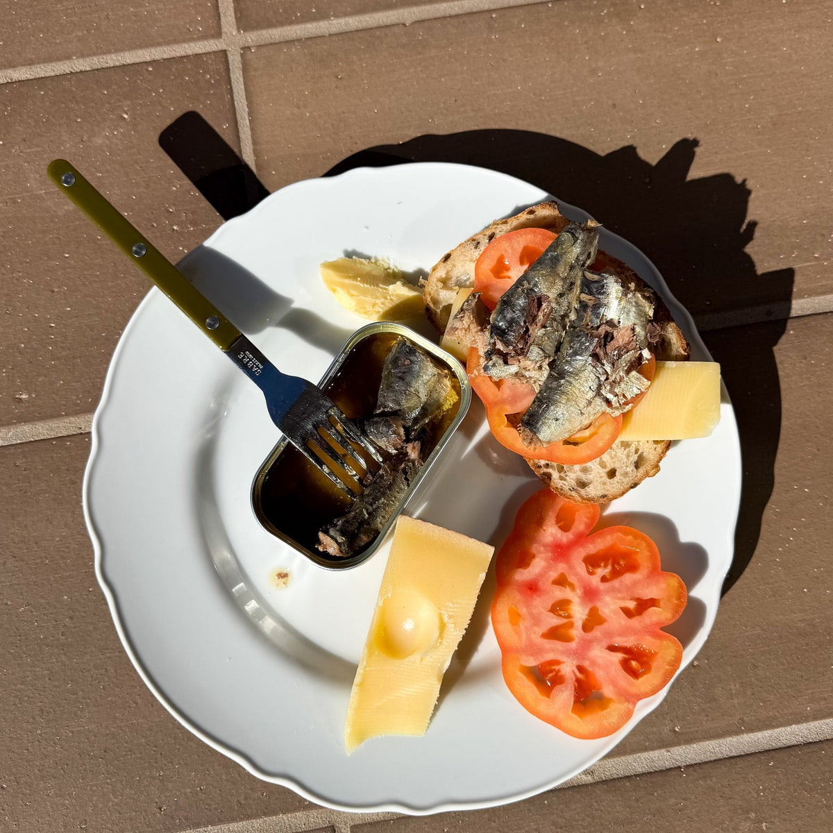 Open tin of smoky style sardines on a plate with tomato and cheese