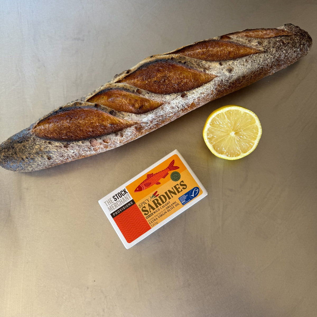 The Spicy Sardines tin on a bench beside a baguette and half a lemon