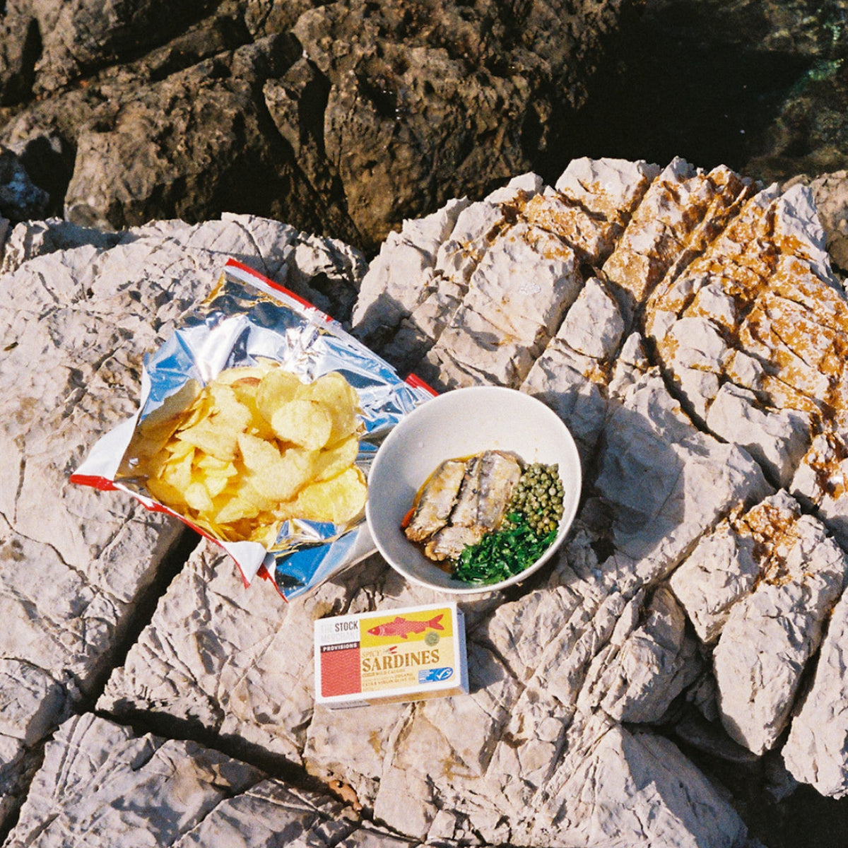 A picnic on coastal rocks with the Spicy Sardines tin, crisps and a bowl of fish and peas
